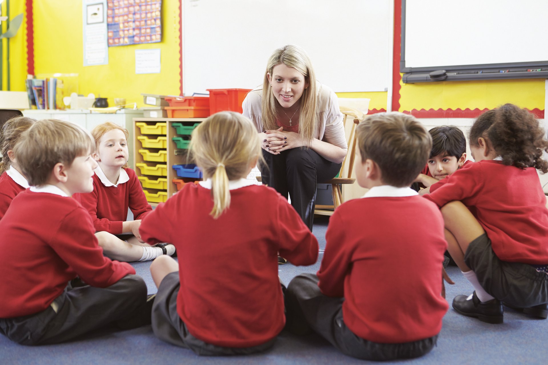 A teacher learning down to a circle of young kids sitting on the floor.