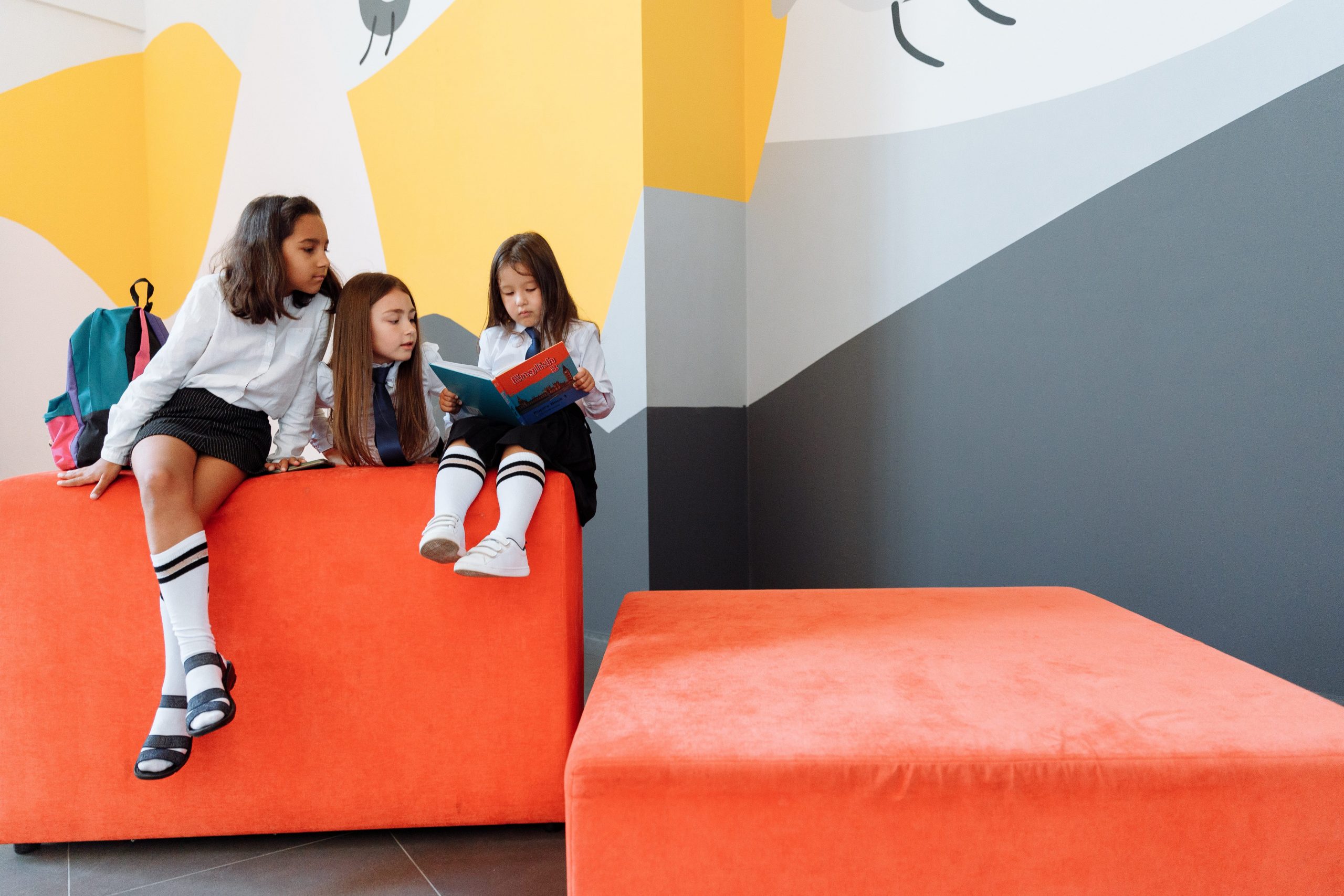 Three girls sitting on a big cubed block all looking at one book.